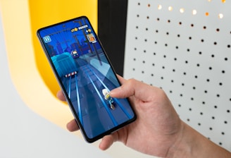 A person is holding a smartphone displaying a mobile game with vibrant graphics. The game depicts a character running on train tracks. The background includes a well-lit wall with a pattern of holes.